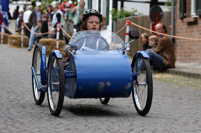 Kinder in flotten Kisten
Der Name Seifenkiste stammt aus den USA: Anfang des 20. Jahrhunderts hatte dort ein Seifenfabrikant seine Holzkisten mit einem Bauplan für so genannte Kinderautos bedruckt und später auch noch passende Achsen und Räder hierzu geliefert. So setze sich der Name Seifenkiste für das Gefährt durch. Seit 1904 werden mit solchen Fahrzeugen auch im deutschsprachigen Raum Wettfahrten auf leicht abschüssigen Strecken ausgetragen, seit 1977 eben auch in Liedberg... 
Schlüsselwörter: Seifenkistenrennen Liedberg